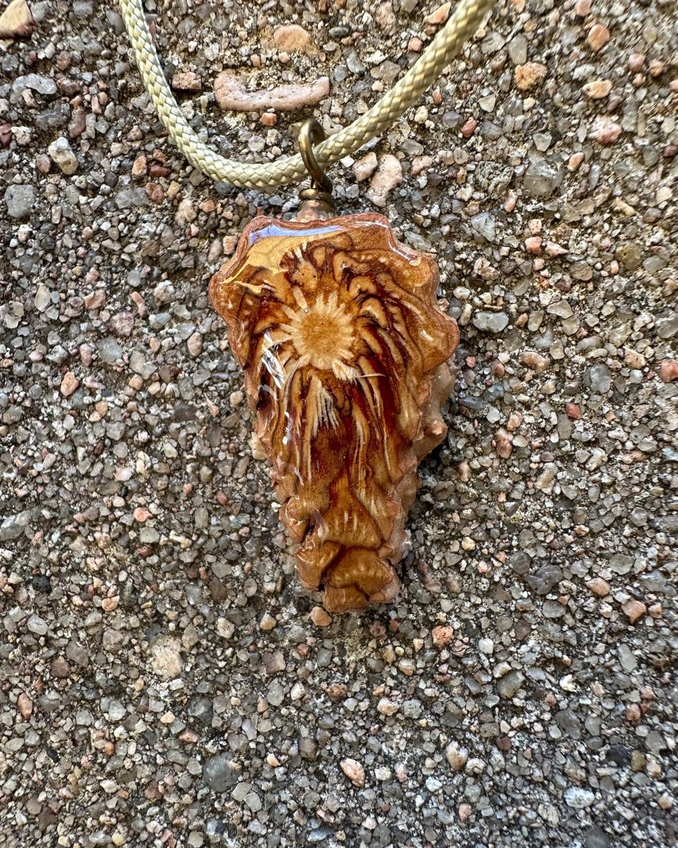 Arrowhead pine cone pendant hanging from a coiled brass loop on a pale braided cord against grey concrete, bright ochre core ringed in cream near the top with rays streaming down to a blunt tip under glossy resin