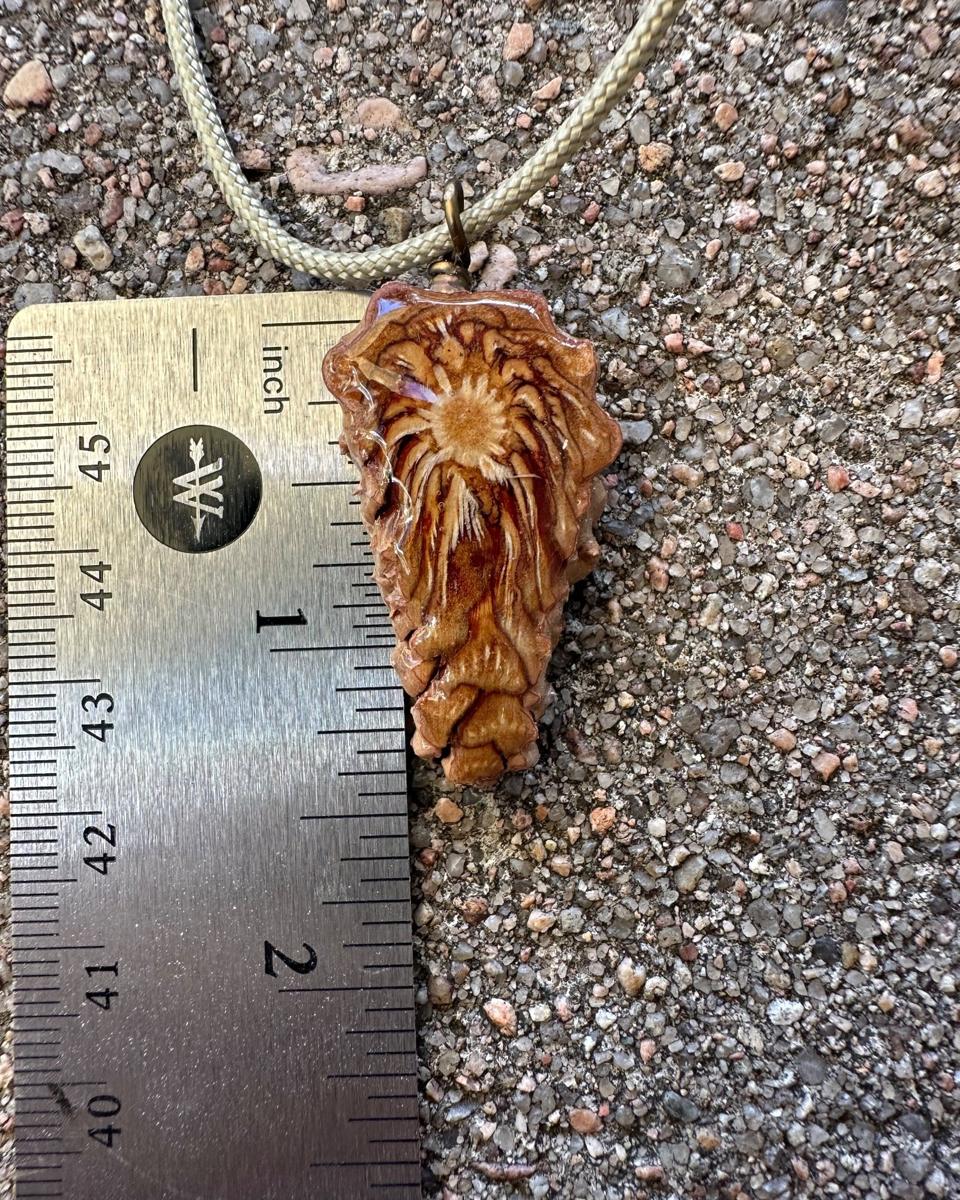 Arrowhead pine cone pendant beside a steel ruler on grey concrete, pale braided cord looping away above it, showing the piece a little over an inch and a half long
