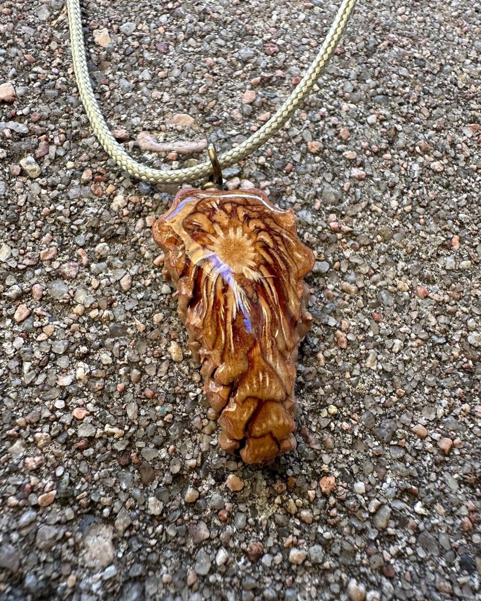 Arrowhead pine cone pendant on a pale braided cord against grey concrete, ochre core near the top with cream and rust rays streaming down and a violet flash across the resin