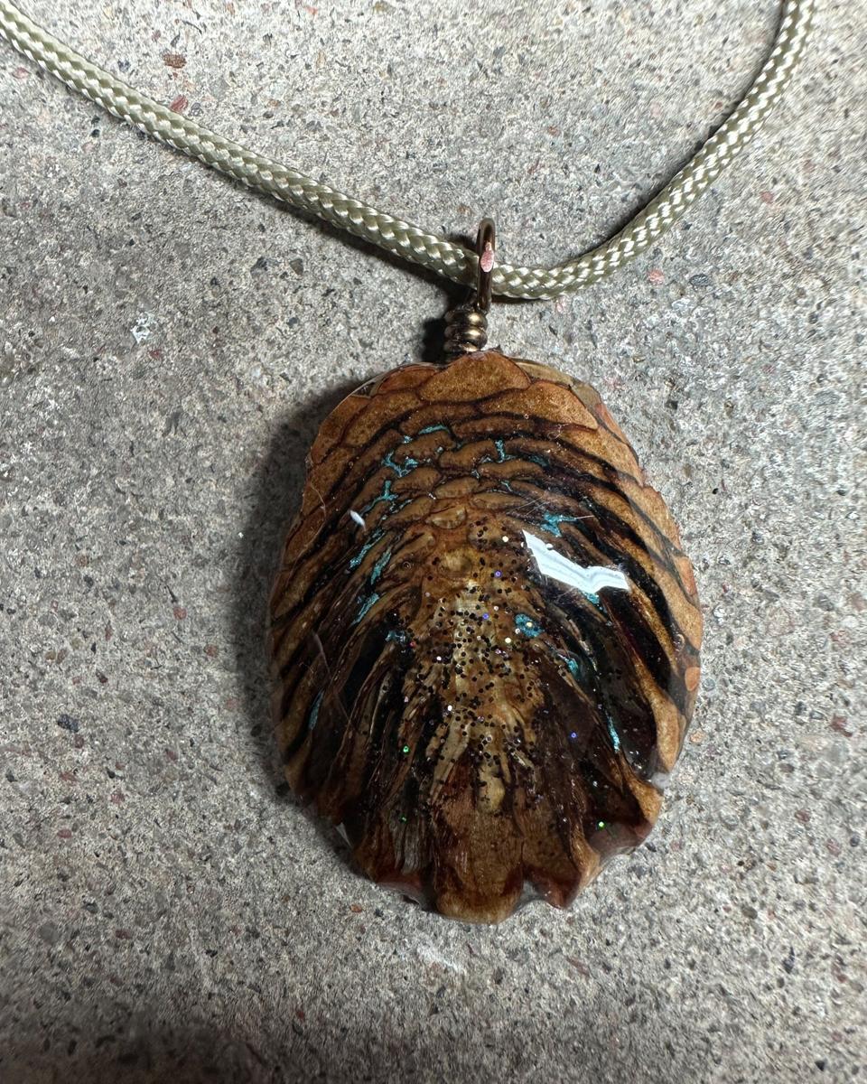 The other face of the pendant, tan scales in stacked rows with turquoise lines in the gaps and dark glitter down the middle, copper loop and pale cord at the top