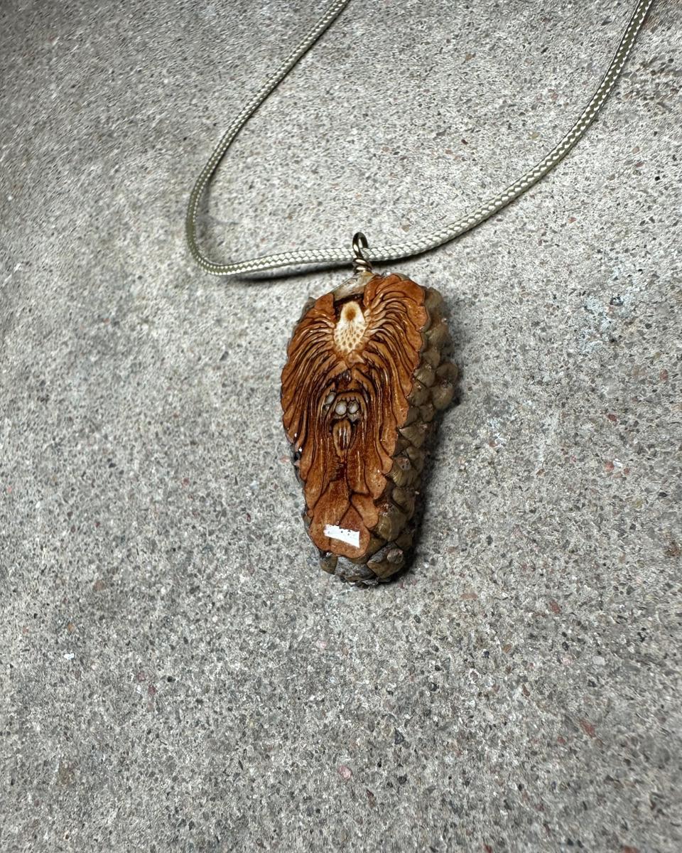 Tall pine cone pendant hanging from a coiled wire loop on a pale braided cord against grey concrete, pale cream core near the top with ochre and rust rays fanning down between dark lines, grey-brown scales along the right side