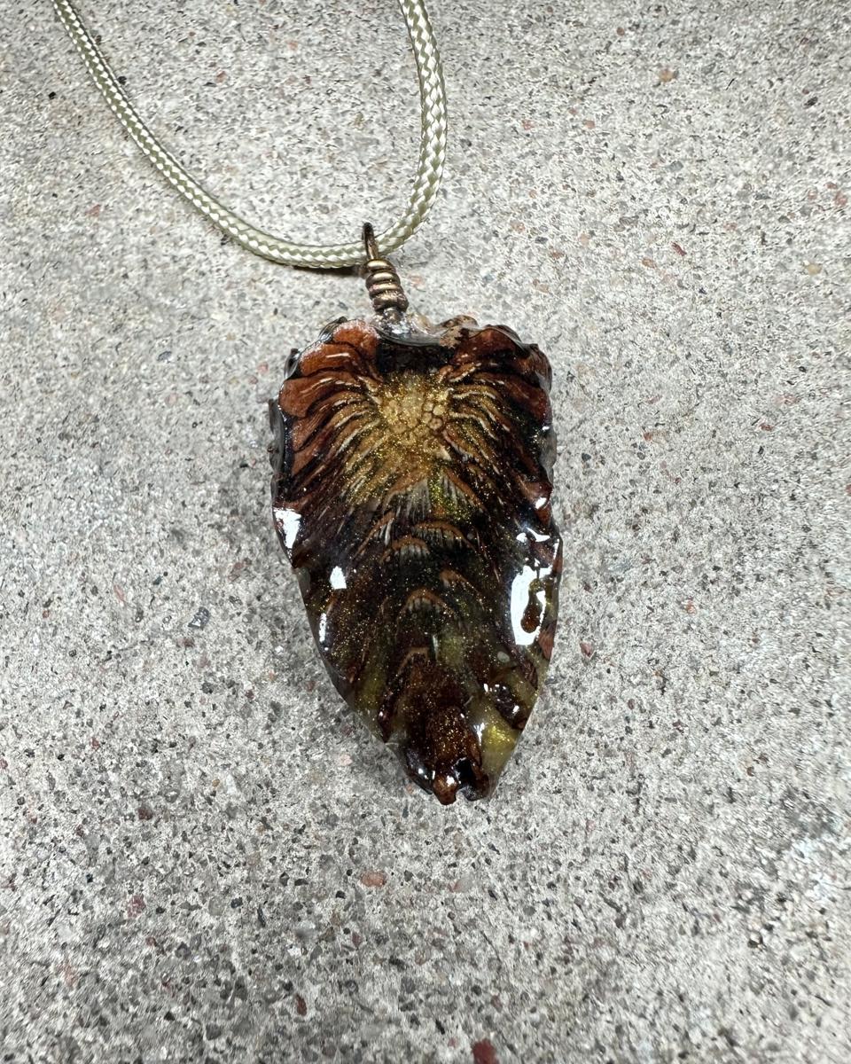 Teardrop pine cone pendant hanging from a coiled wire loop on a pale braided cord against grey concrete, pale gold burst near the top with tan rays fanning down into olive and near-black under glossy resin