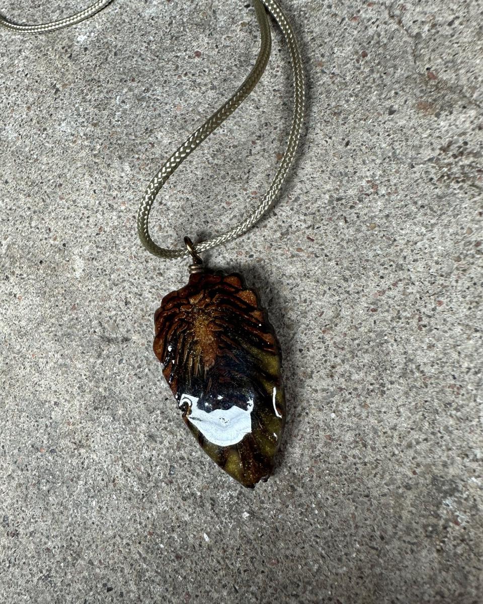 The other face of the pendant on its pale cord, rust scales at the top fanning down into dark olive with a white highlight across the lower half