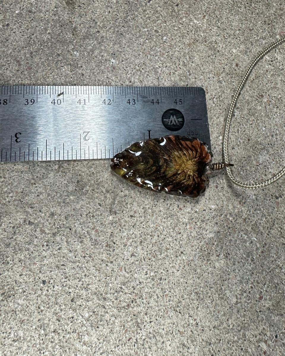 The pendant beside a steel ruler on concrete, cord looping away to the right, showing the teardrop outline and gold burst
