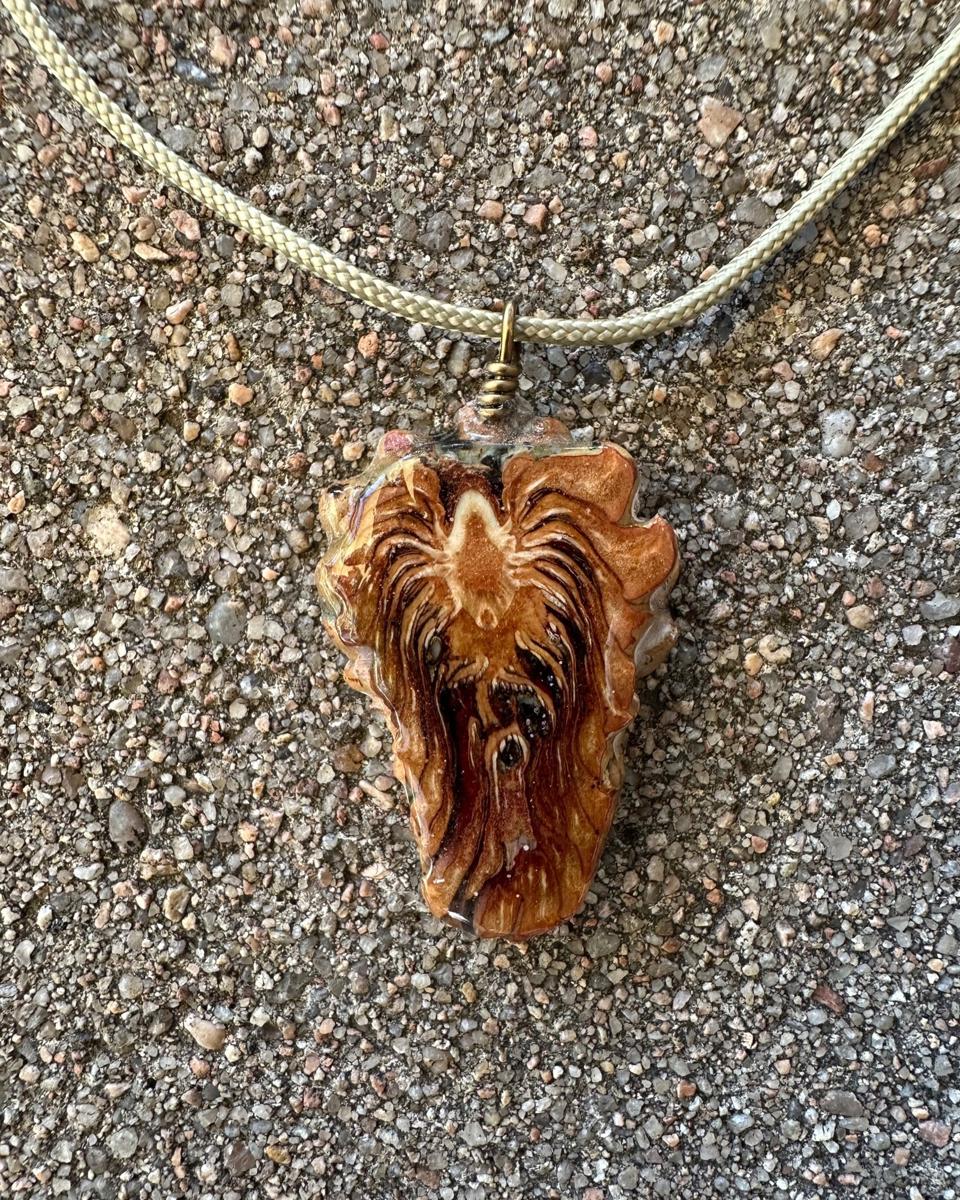 Spade-shaped pine cone pendant hanging from a coiled brass loop on a pale braided cord against grey concrete, pale cream core near the top with cream and rust rays spraying down into a dark red-brown centre under glossy resin