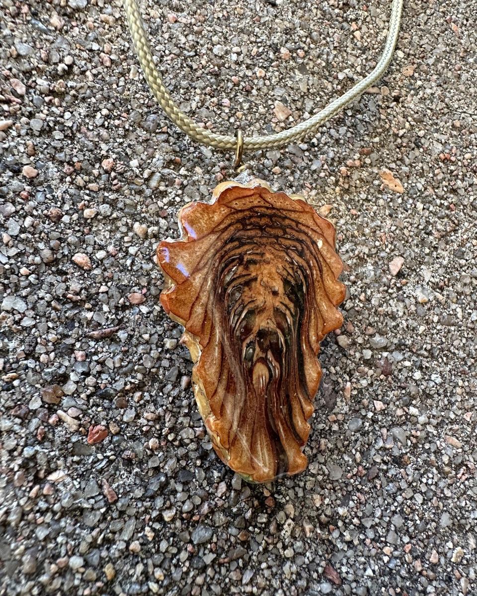 The other face of the pendant on its pale braided cord, rust and honey scales in stacked arcs with dark brown lines running down the middle to a blunt tip