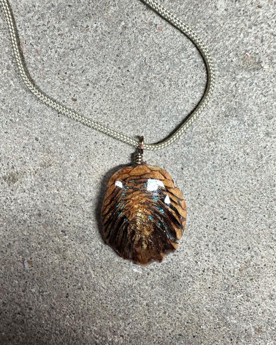 Oval pine cone pendant hanging from a copper loop on a pale braided cord against grey concrete, tan and rust scales with turquoise veins and a glittered center