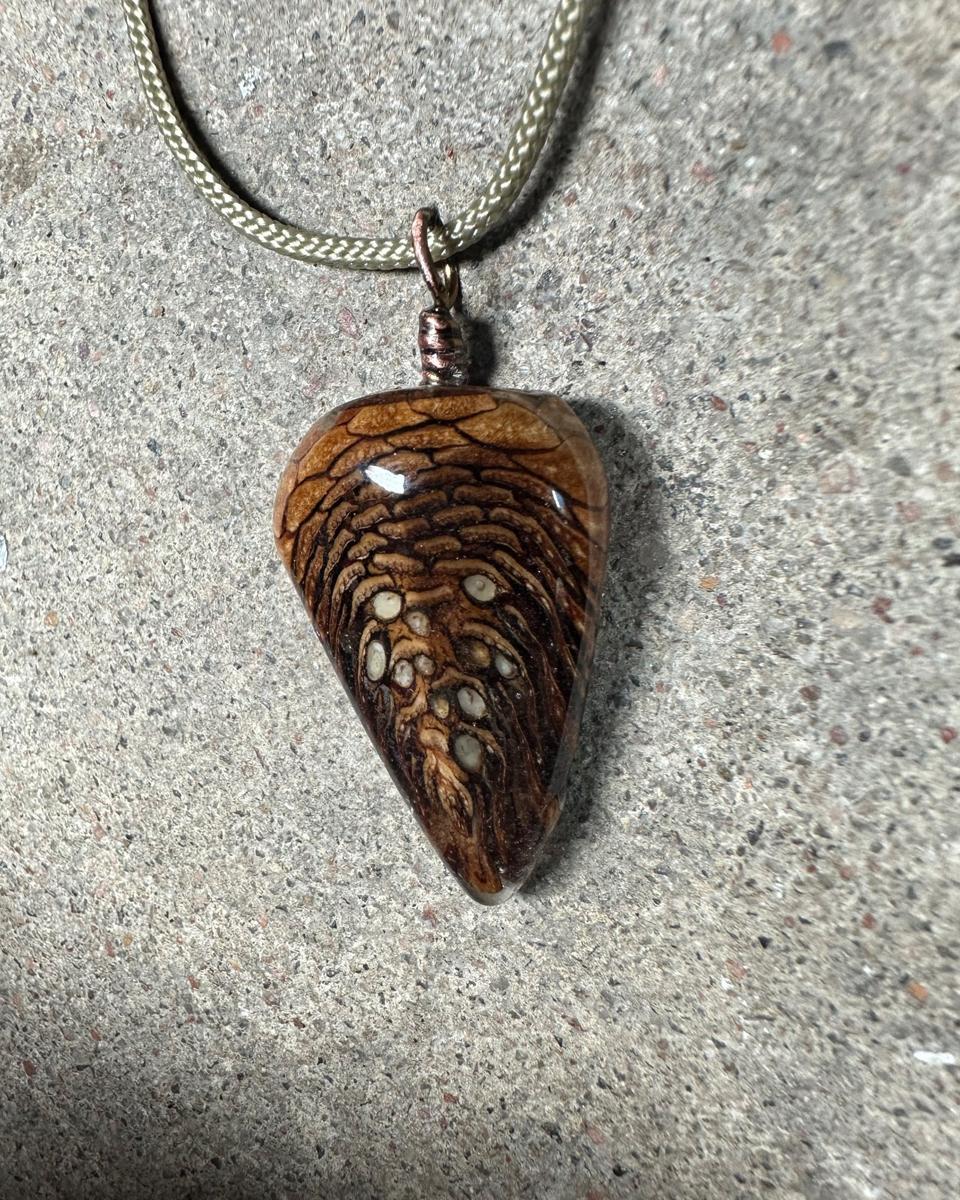 The pendant hanging from a copper loop on a pale braided cord, scale rows in tan and rust stacking down to a dark point with pale seed ends near the bottom