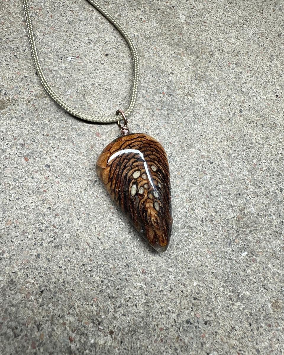 Rounded triangle pine cone pendant lying on grey concrete with a pale braided cord looping away, tan scales at the top grading into dark brown, glossy resin catching a white highlight