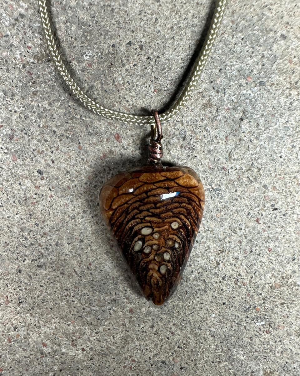 Closer view of the pendant on its cord, showing the flat tan facets at the top, dark lines between the scales and cream seed ends low on the face