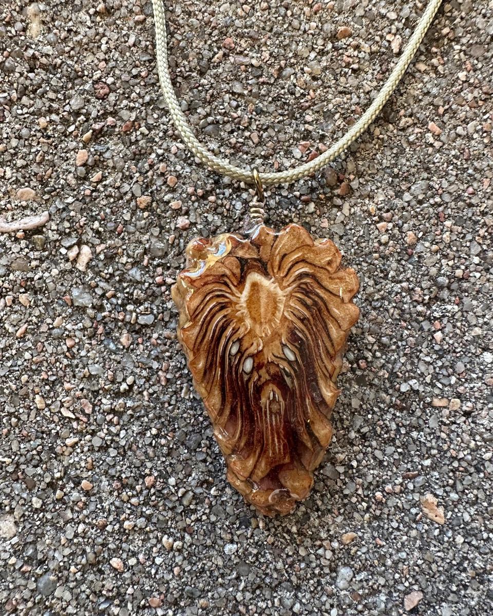 Arrowhead pine cone pendant hanging from a coiled wire loop on a pale braided cord against grey concrete, pale core near the top with rays streaming down to a blunt tip under glossy resin