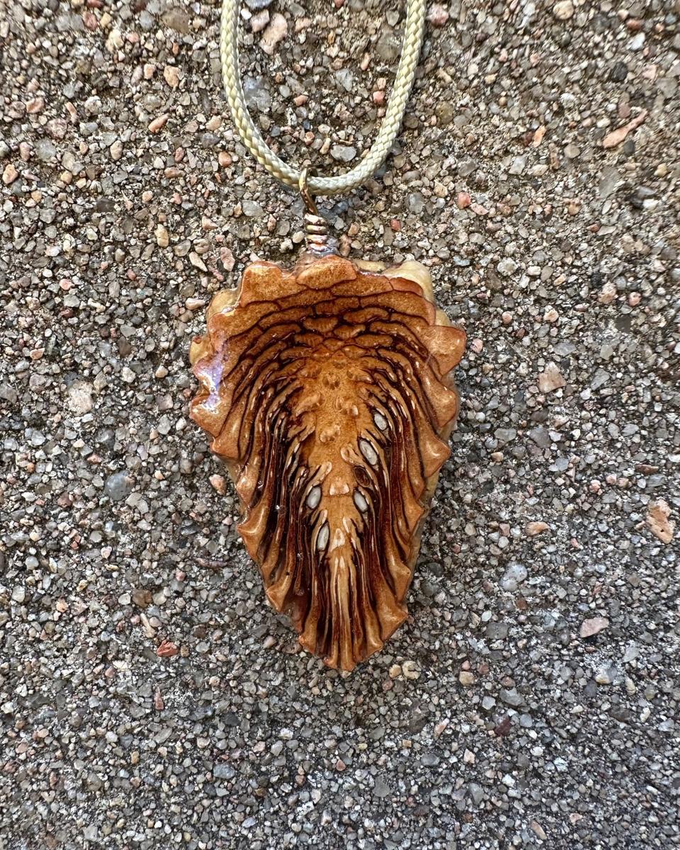 Arrowhead pine cone pendant on a pale braided cord against grey concrete, dark brown rays streaming down from a ridged top into a tan and rust tip