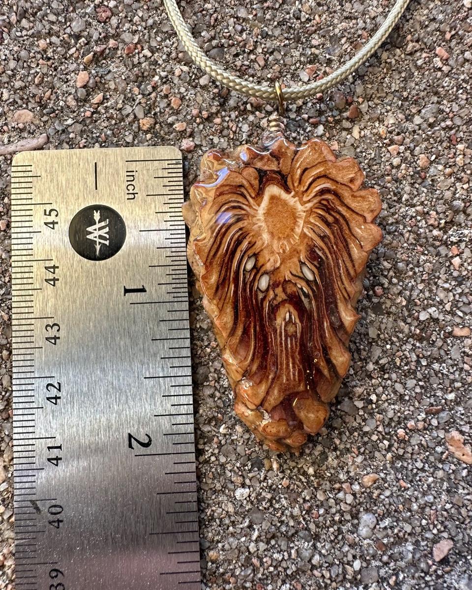 The pendant beside a steel ruler on concrete, cord looping away above it, showing the arrowhead outline a little over two inches long
