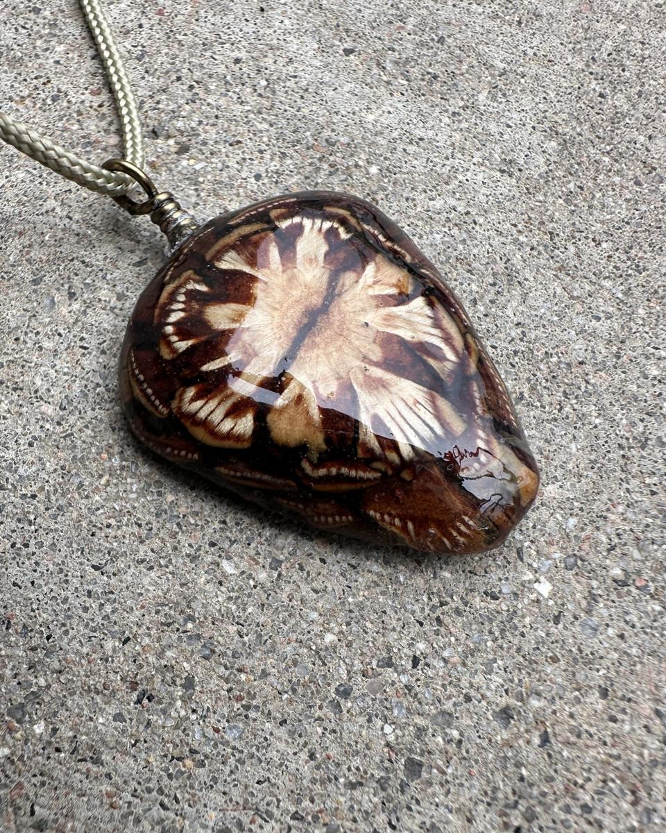Shield-shaped dark brown pendant with a cream winged burst at its center under a domed resin surface