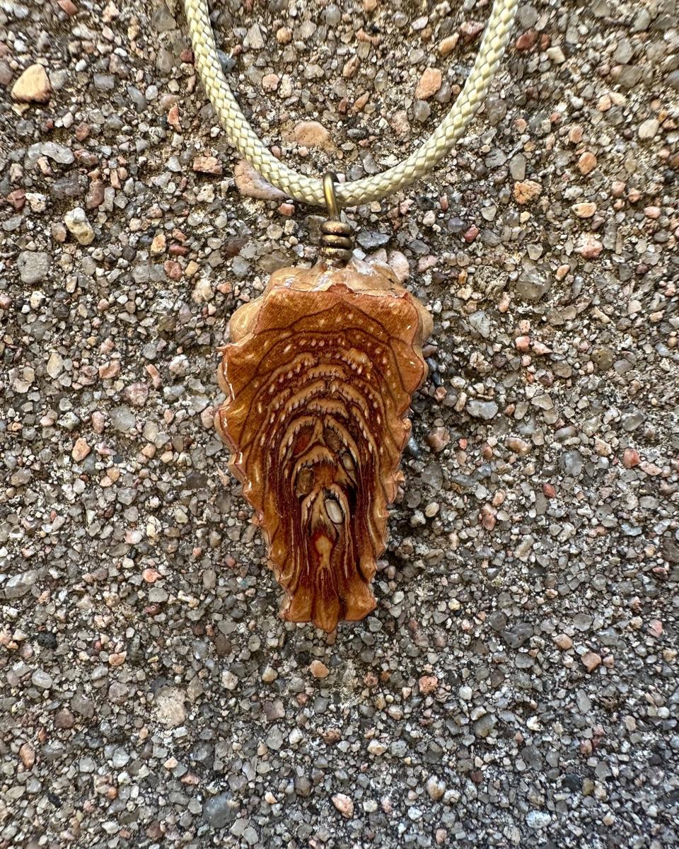 The other face of the pendant on its pale braided cord, rust and honey scales stacked in arcs with dark brown lines between the rows and small white flecks along them