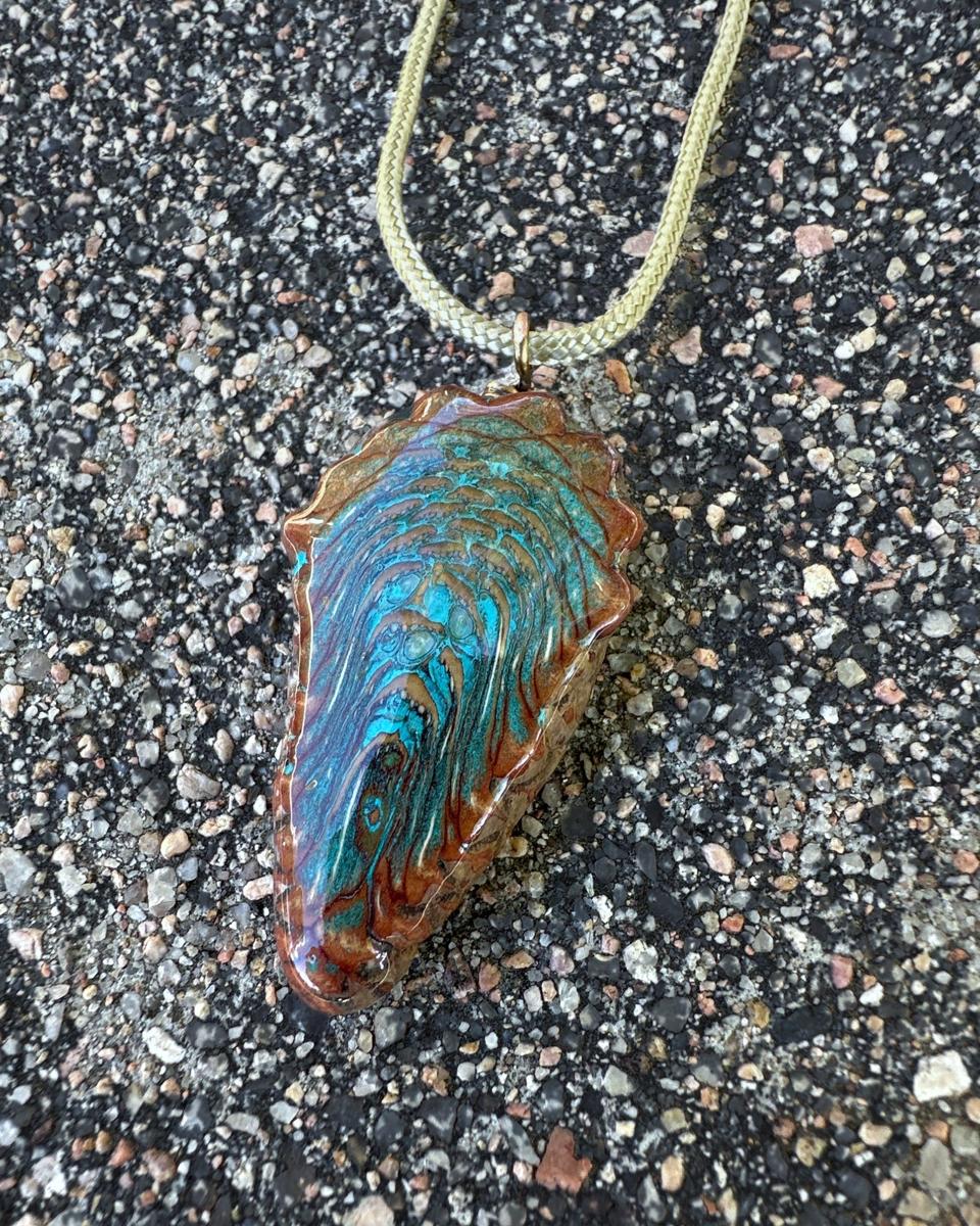 Long teardrop pine cone pendant in turquoise and rust, hanging from a pale braided cord on grey gravel