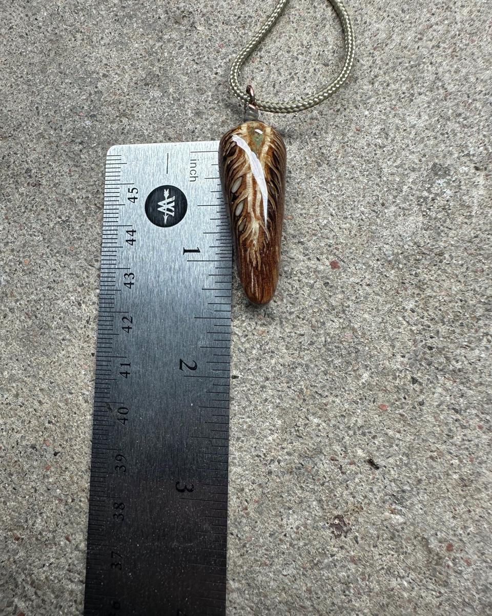 The pendant beside a steel ruler on concrete, cord looping away above it, showing the long teardrop outline