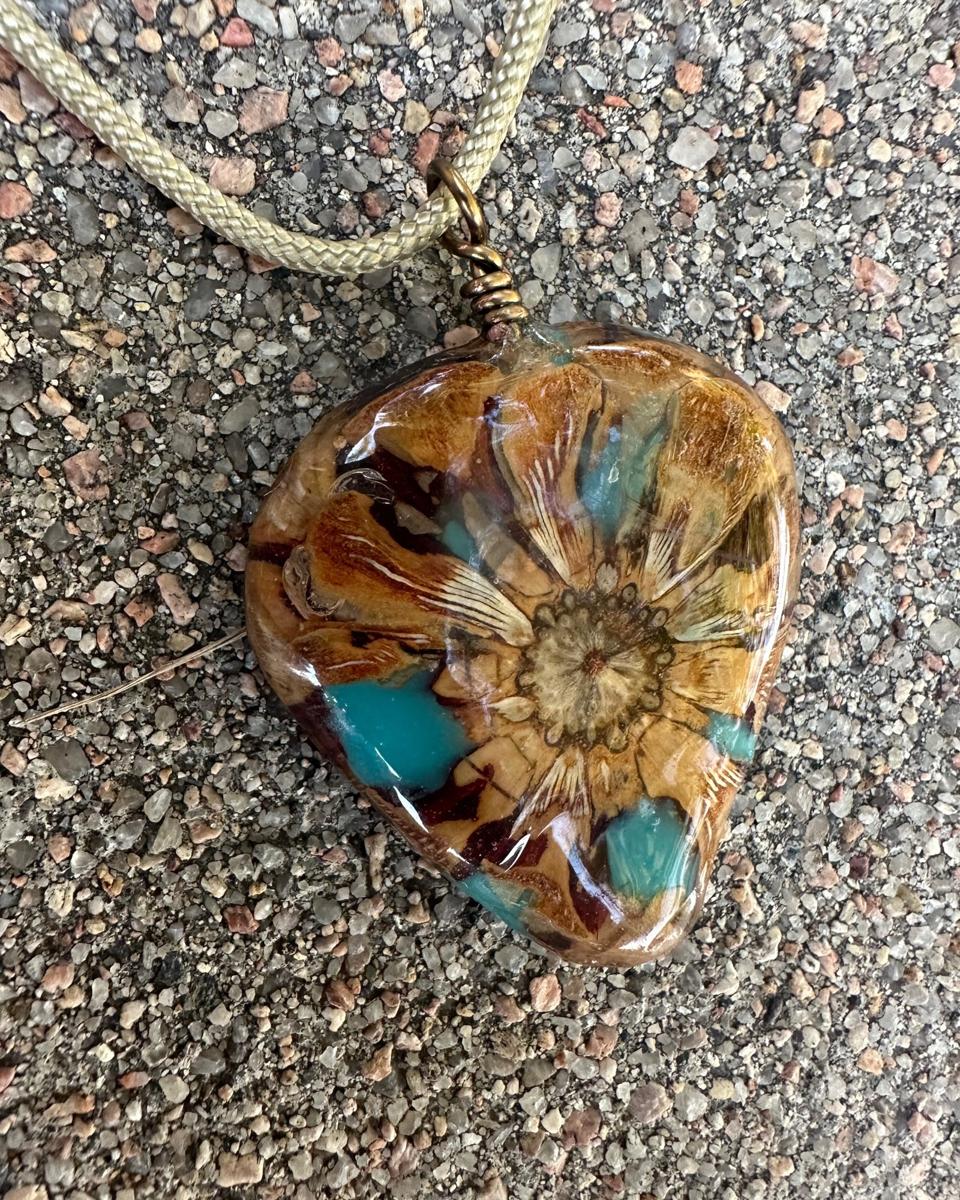 Rounded triangle pine cone pendant on a pale braided cord, tan and rust scales fanning out from a round cream rosette, with turquoise and dark red patches under glossy resin