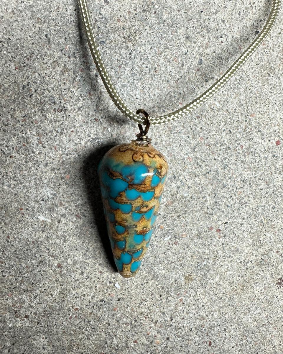 The pendant hanging on a pale braided cord, whole cone shape tapering to a rounded tip, tan and olive scales with turquoise spots, casting a shadow on grey concrete