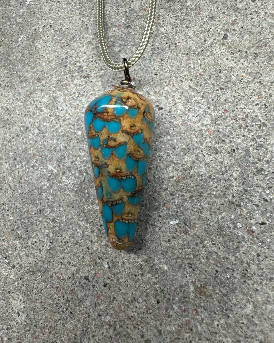 Long cone pine cone pendant hanging from a pale braided cord against grey concrete, tan scales with turquoise drops in the gaps under glossy resin