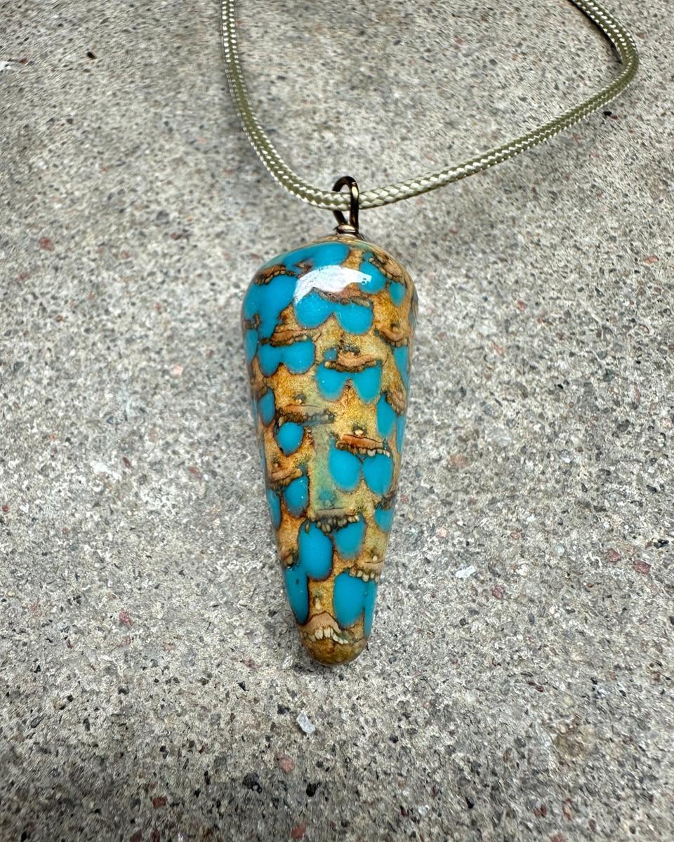 Closer view of the pendant on its cord, staggered rows of tan scales with bright turquoise drops between them and a white highlight near the top