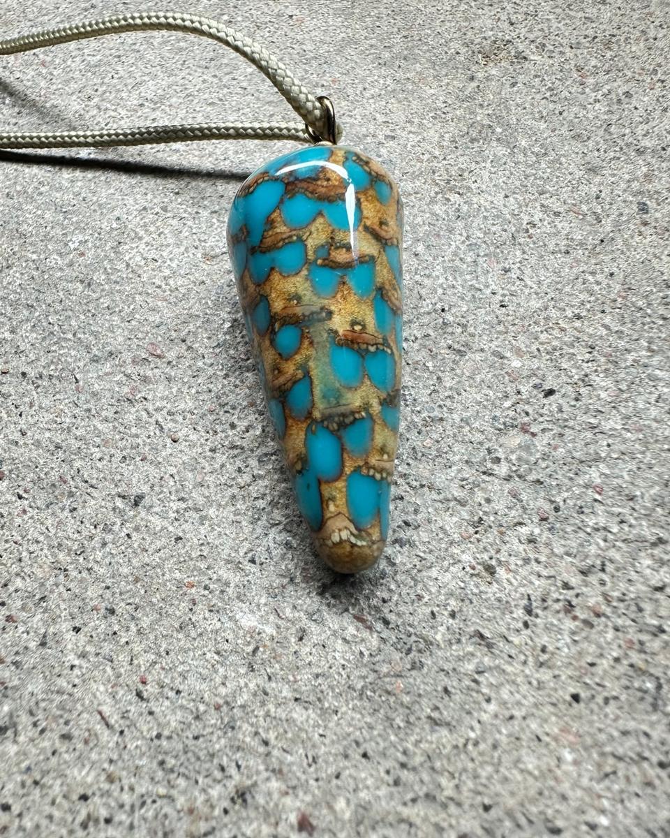 The pendant lying at an angle with the cord looping away, showing the tapered tip and turquoise spots running down the tan scales