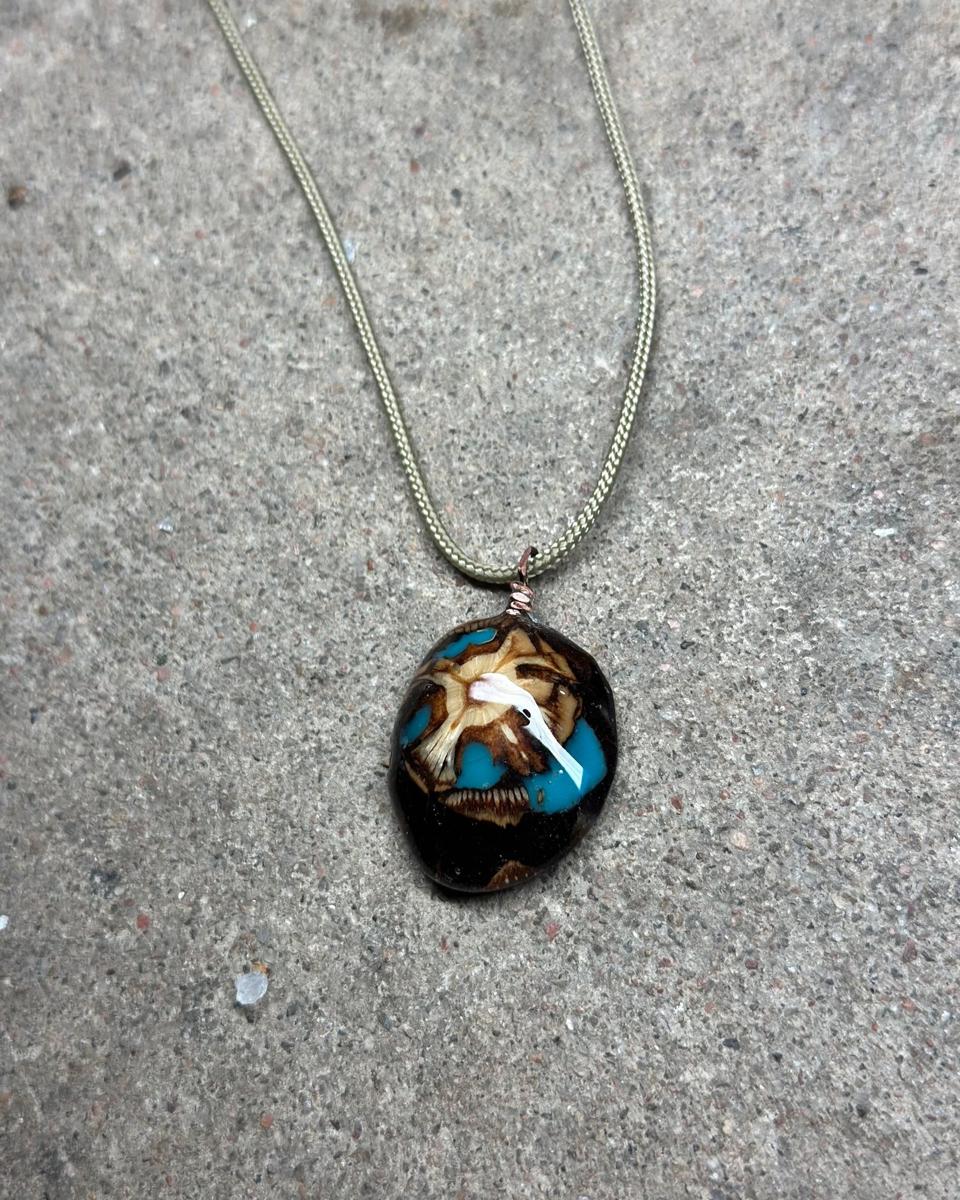 Oval pine cone pendant on a pale braided cord against grey concrete, dark face with a cream star at the center and turquoise pools between the scales