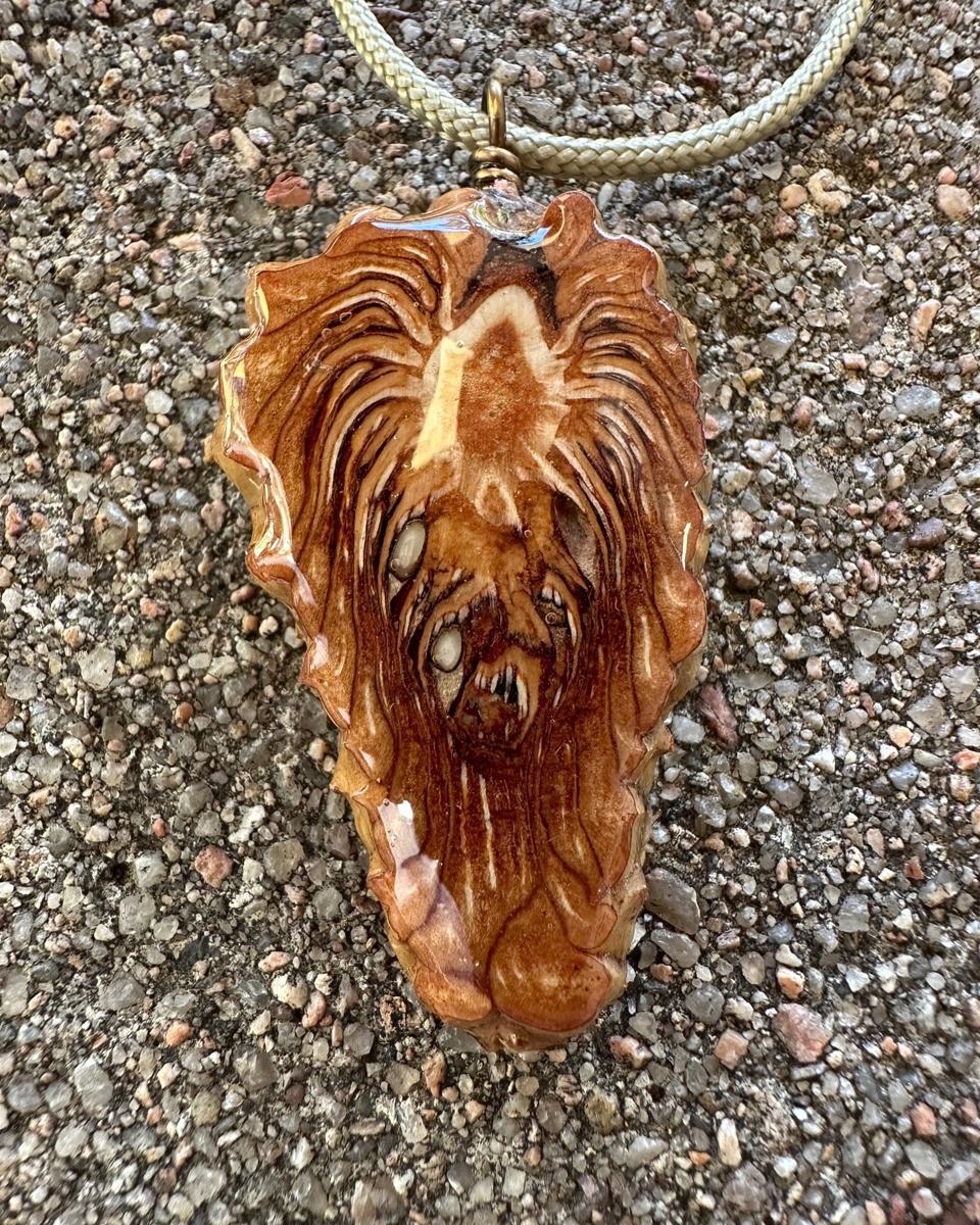 Arrowhead pine cone pendant hanging from a brass loop on a pale braided cord against grey concrete, cream patch near the top with long rust and brown strands parting down the middle under glossy resin