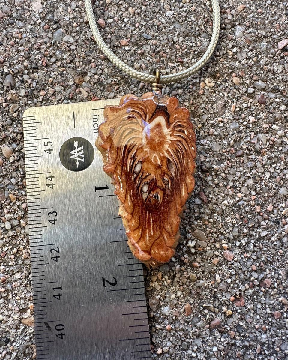 Arrowhead pine cone pendant beside a steel ruler on grey concrete, pale braided cord looping away above it, showing the piece a little over an inch and a half long