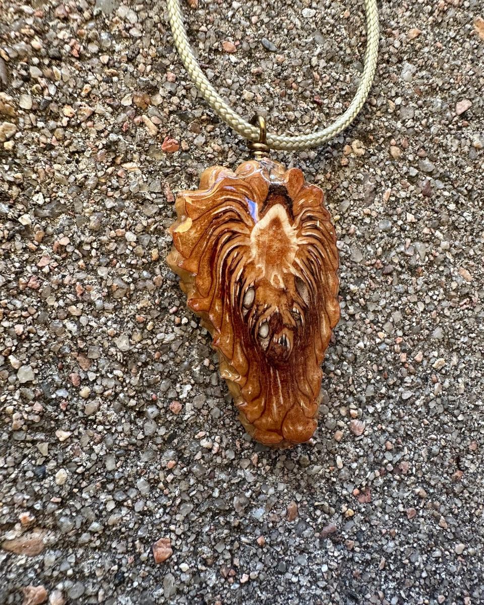 Arrowhead pine cone pendant on a pale braided cord against grey concrete, pale cream patch near the top with rust strands falling to a blunt tip under glossy resin