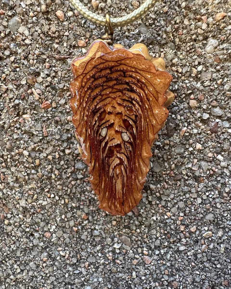 The other face of the pendant on its pale cord, rust and tan scales in stacked arcs with pale flecks along the ridges and long strands running to the bottom